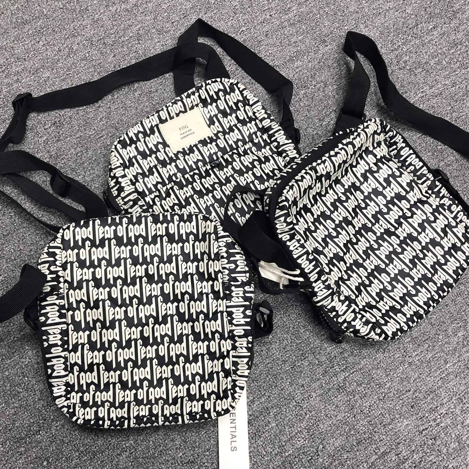 Black and white printed tote bag and backpack featuring the repeated text "fear of god" on a gray carpeted floor.