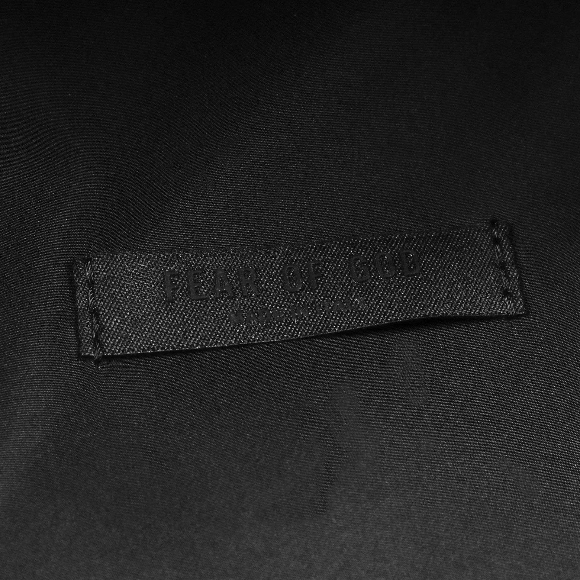 Black fabric tag with "FEAR OF GOD" embossed, sewn onto a dark material, indicating high-end fashion merchandise, possibly related to K-pop inspired streetwear or luxury K-pop merchandise collections.