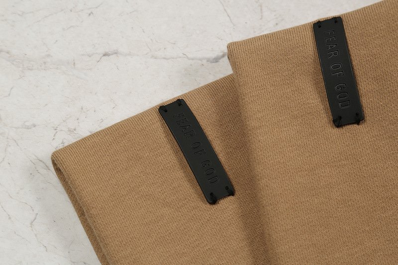 Black rubber tags with "FEAR OF GOD" embossed, attached to tan fabric, showcasing a trendy K-pop merchandise design.