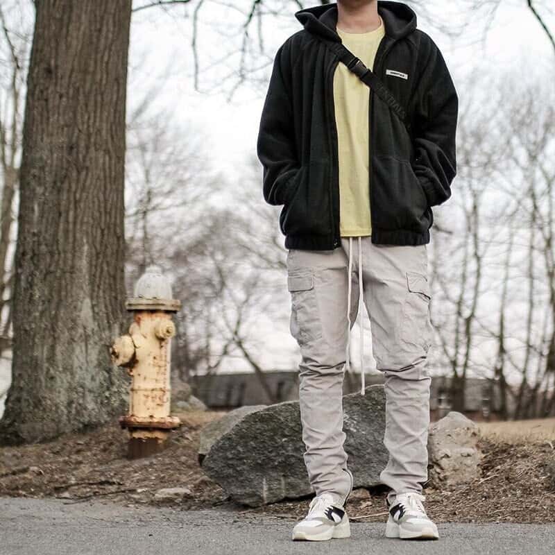 Fashion-forward casual hoodie and cargo pants outfit for men on a fall day outdoors, with a person standing in a park-like setting featuring trees and bare branches in the background, trendy streetwear style, comfortable and stylish look, popular among K-pop fans and merchandise enthusiasts.