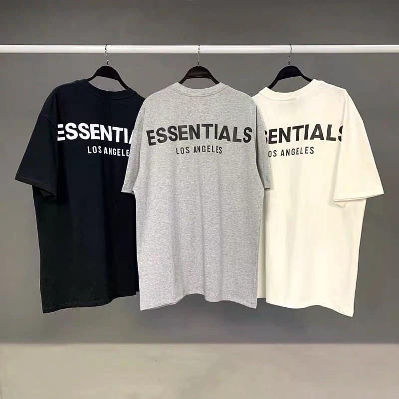 ESSENTIALS Los Angeles graphic T-shirts in black, gray, and white, laid flat hanging on black hangers against a grey background. Trendy K-pop merch emphasizing minimalist streetwear style.